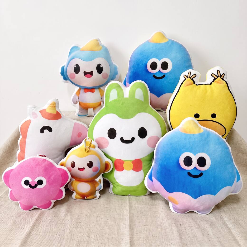 Eight shaped plush pillows on a linen cloth: a green bunny, a yellow duck, a unicorn, a pink blob, two round blue birds and more.